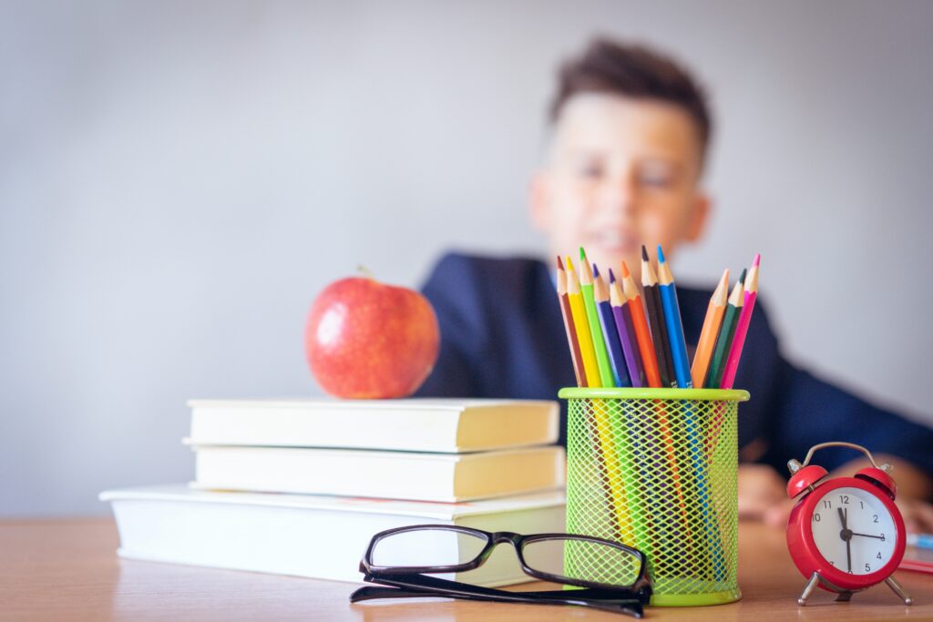 A child sits behind a table with books, colored pencils, an apple, glasses and a clock, preparing to go back to school.